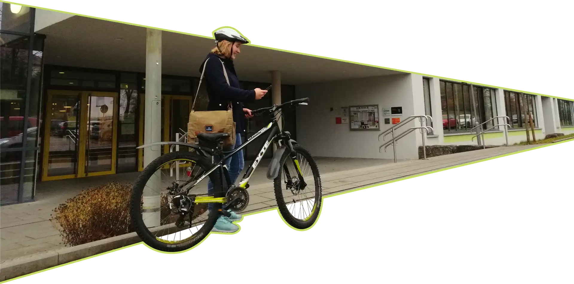 Eine Schülerin trägt am Ziel angekommen ihre zurückgelegte Strecke ein. Schülerin steht vor Schule, angelehnt an ihr Fahrrad und benutzt ihr Handy.
